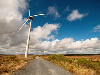A field of wind turbines with a clear blue cloudy sky in the background. The windmills are tall and...