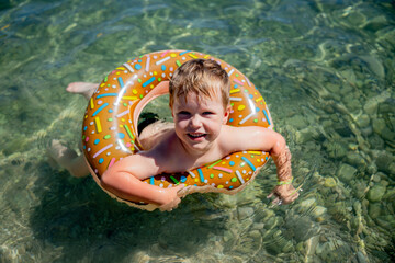 A little happy boy bathes in a doughnut lifebuoy in clear sea in sunny weather