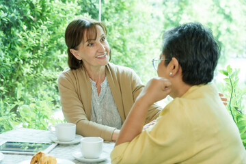 Asian senior woman and Caucasian woman sit at a cafe table, smiling warmly as they engage in a meaningful conversation over tea and pastries, enjoying a joyful retirement moment together.