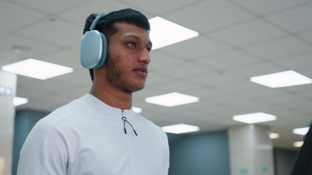 young man clips over ear headset onto neck steps onto treadmill powers console and begins cardio session in spacious gym lit by overhead panels with equipment and exerciser blurred in background