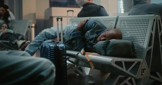 Crowded International Airport Terminal: Exhausted African American Man with Luggage Sleeping on the Seats in Departure Lounge of Airline Hub. Diverse People and Passengers Waiting for Delayed Flight.