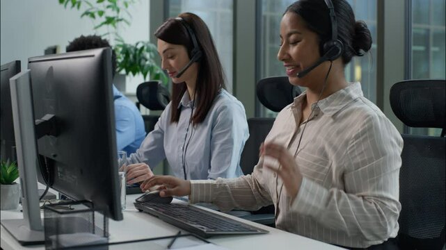 Three business people multiethnic Indian Caucasian women African man headset call center service operators work indoor callcenter talk client using pc computer writing notes helpful customer service