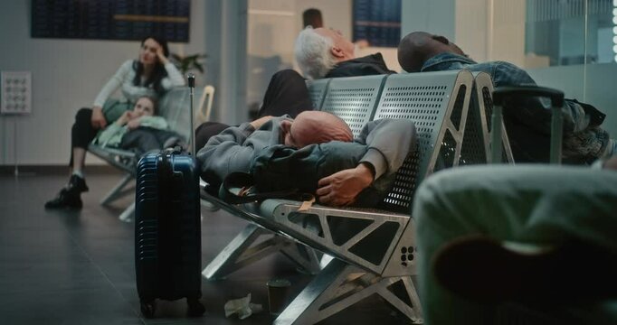 Crowded International Airport Terminal: Exhausted Man with Luggage Sleeping on the Seats in Boarding Lounge of Airline Hub. Diverse People and Passengers Waiting for Delayed Flight. Dolly Shot Zoom In