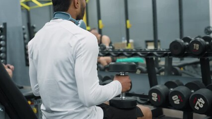 side view of gym rat in white shirt sitting and placing dumbbell on leg inside well equipped gym with visible dumbbells and racks, another gym-goer passes by in background