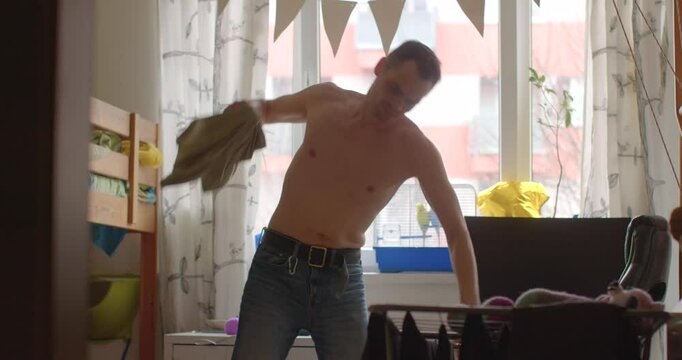 A man is changing his clothes in a cozy bedroom during the daylight hours, with a soft background ambience