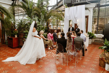 The bride walks towards the groom and priest who are standing opposite her while a group of nine...