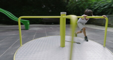 Boy spinning on a playground carousel, surrounded by green trees and a wooden fence in a serene park setting