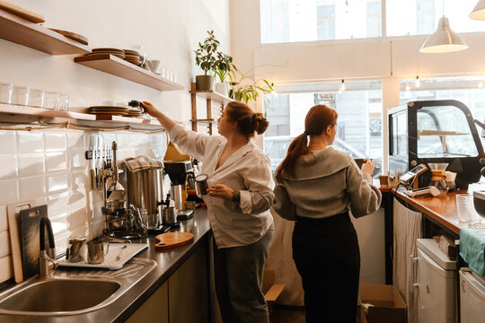 A female owner stands behind the bar and displays teapots on a shelf while a female employee stands next to her, in a cafe