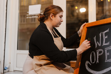 The female owner sits and writes with chalk on a sign, on the street at the entrance to the cafe