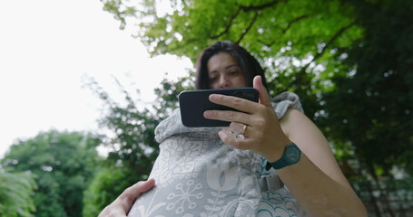 Woman multitasking in a park, holding her baby in a front carrier while focused on her smartphone,...