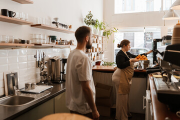 A male employee stands behind the bar and looks at the female owner who stands in front of him and...