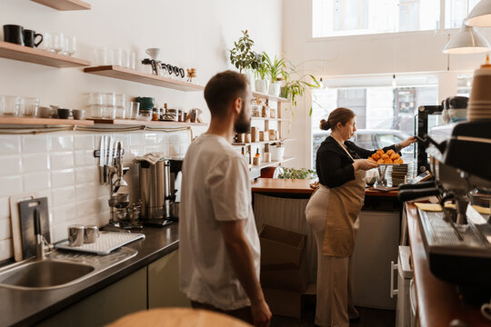 A male employee stands behind the bar and looks at the female owner who stands in front of him and puts profiteroles on the display case, in a cafe