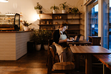 The female owner is sitting at a table with a laptop and writing with a pen in a notebook, in a cafe