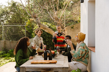 Group of five friends talking and holding glasses while sitting and standing at a table, outdoors