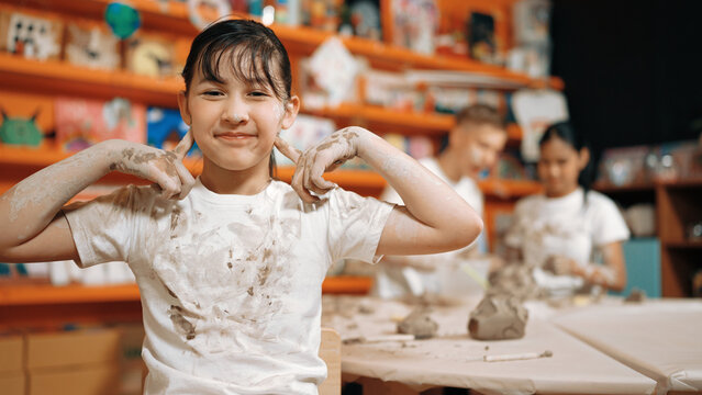 Asian girl pose at camera while diverse children modeling clay behind. Happy cute student wearing dirty shirt while looking at camera at workshop in art lesson. Blurring background. Edification. - Powered by Adobe