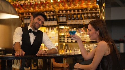 Sitting in smiling beautiful woman guest lively taking with smart bartender while drinking mixed vodka in blue hawaii cocktail and whiskey at luxurious counter bar on Friday meeting at club. Vinosity.