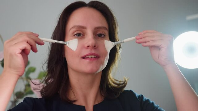 portrait view of cheerful female cosmetologist teasingly brushing cheeks with twin silicone facial brushes under ring light in modern bright spa room with floral decor and skincare products