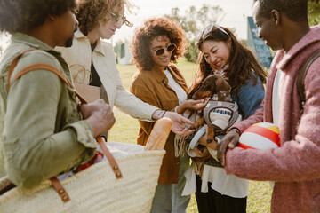 A group of five friends stand and laugh while two of them pet a dog held by another, outdoors