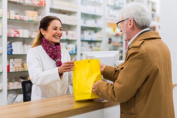 A friendly pharmacist gives a yellow prescription bag to an elderly man in a pharmacy.