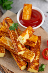 Fried halloumi cheese sticks with tomato sauce on a wooden board on a concrete background.