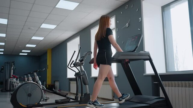 sport woman keeps fit walking on treadmill under sunlight through large window in modern gym with wall clock on gray wall row of exercise bikes and elliptical trainer visible in background