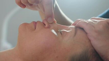 Bodywork expert massages client nose with gentle fingertip movements in calm spa environment, applying smooth pressure along nasal bridge to release tension and ease stress under ambient lighting