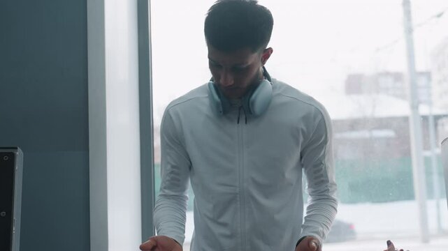 male fitness trainer in white jacket and headphones monitors bench press form as female student lifts barbell indoors, with snow-covered street and passing cars visible through large window