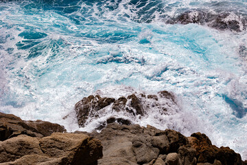 Turquoise ocean waves swirling and crashing against rugged coastal rocks in bright sunlight 