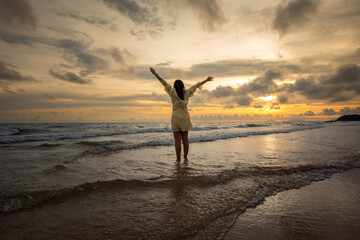 Beautiful Asian woman walking peacefully on a desert beach during sunset, with freedom concept, with amazing orange sky and sunlight reflecting on the sea and sand.