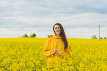 Young beautiful woman in a yellow hoodie close up in the middle of the yellow field with rapeseed flowers on the blue sky background. Republic of Belarus, Brest region