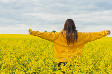 Young beautiful woman in a yellow hoodie close up in the middle of the yellow field with rapeseed flowers on the blue sky background. Republic of Belarus, Brest region