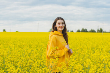 Young beautiful woman in a yellow hoodie close up in the middle of the yellow field with rapeseed...