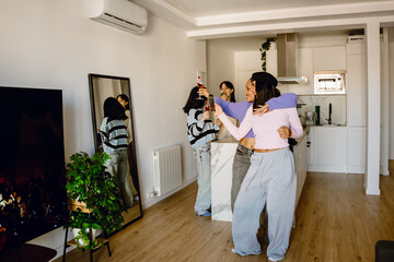 Two female friends standing and smiling while posing in front of a mirror while one of them holds a phone and the other a glass