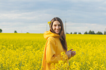 Young beautiful woman in a yellow hoodie close up in the middle of the yellow field with rapeseed flowers on the blue sky background. Republic of Belarus, Brest region
