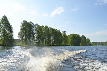 Scenic view of a calm river with lush green trees along the banks from a motorboat leaving a wake on the water. Concept of traveling on water by motorboat on a summer day