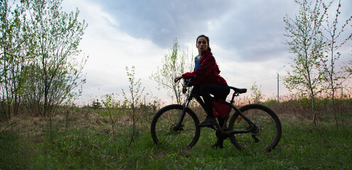 Fototapeta premium Young woman riding a bicycle on the spring field with green grass on sunset sky landscape background. Happy activity sport trip vacations
