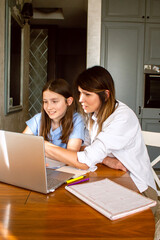 Mother and daughter using laptop sitting together in home kitchen. Mom helping kid with school...
