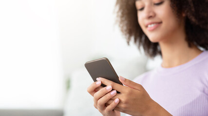 Fototapeta premium Modern online communication during covid-19 lockdown, home alone, new normal, be safe. Cheerful young african american woman typing message or surfing on phone in living room interior, selective focus