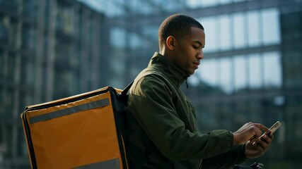 African american man courier checking order on smartphone while riding bicycle with thermal bag. Fast delivery service concept. Urban cityscape on backdrop.