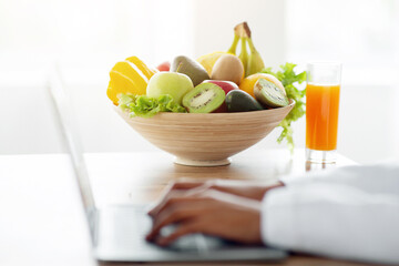 Dietology and healthy nutrition concept. Weight loss consultant working on laptop, giving online consultation to patient at clinic, focus on bowl of fruits and veggies with glass of juice
