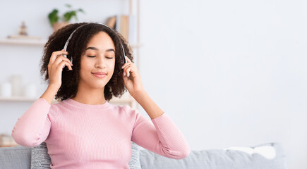 Leisure time, weekends and relaxation with music. Smiling millennial african american woman with closed eyes putting on wireless headphones sitting on sofa and enjoying song in living room interior