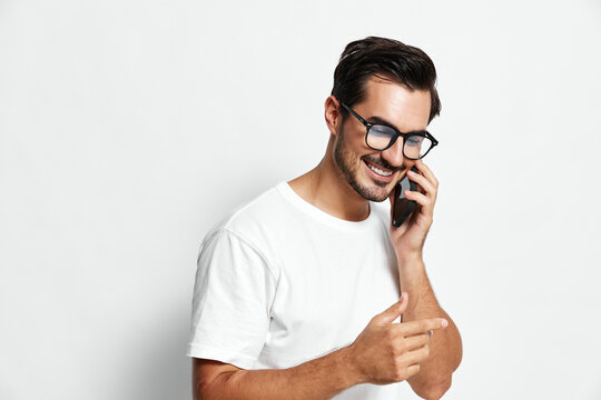 Happy smiling man talking on mobile phone while pointing at something, wearing casual white t-shirt, isolated on plain solid background for lifestyle portrait. - Powered by Adobe