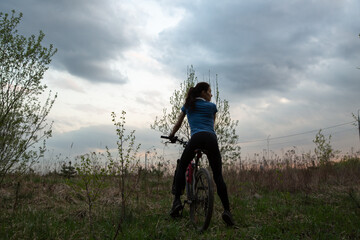 Young sporty woman riding a bicycle on the spring field with green grass on sunset sky landscape...