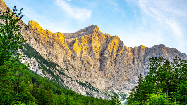 Fototapeta Triglav Mountain stands tall in the Julian Alps, bathed in golden light during sunset. Lush greenery at its base highlights the rugged texture of the rock faces.