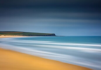 Coastal landscape with a dramatic sky and gentle waves.  A sandy beach meets a cliff, and the water displays a soft, flowing motion