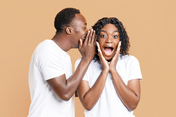 African-american man sharing secret or whispering gossips into his girlfriend's ear, yellow studio background. Black guy whispering words of love to his amazed woman who screaming and touching cheeks