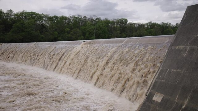 Slow motion wide landscape of a concrete dam holding and overflowing with water, waves, bubbles, minerals, and industrial equipment after recent rainfall, causing a strong stream and crashing liquid