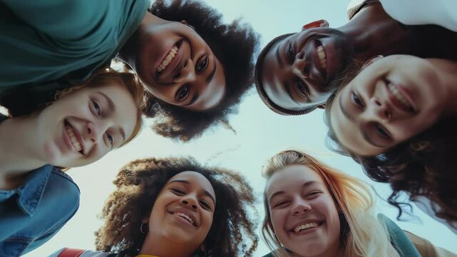 A diverse group of young women and men are shown closely gathered together, smiling brightly as they look down into the camera from a low angle, in a bright and cheerful atmosphere.