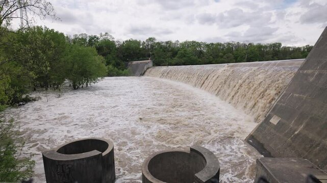 Slow motion wide landscape of a concrete dam holding and overflowing with water, waves, bubbles, minerals, and industrial equipment after recent rainfall, causing a strong stream and crashing liquid