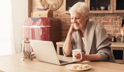 Happy Aged Woman Relaxing In Kitchen With Laptop And Coffee, Watchig Videos Or Browsing Internet, Panorama With Free Space